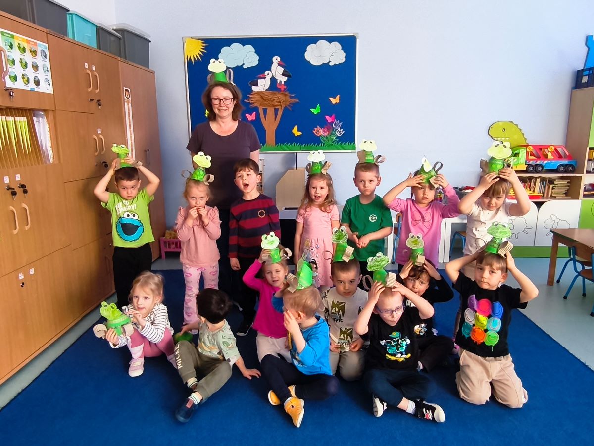 grupa dzieci przedszkolnych stoi razem w przedszkolnej Sali edukacyjnej i prezentuje wykonane przez siebie papierowe żabki. Dzieciom towarzyszy edukatorka ekologiczna i nauczycielka przedszkolna. Dzieci trzymają wykonane z papieru i tektury żabki na głowie.