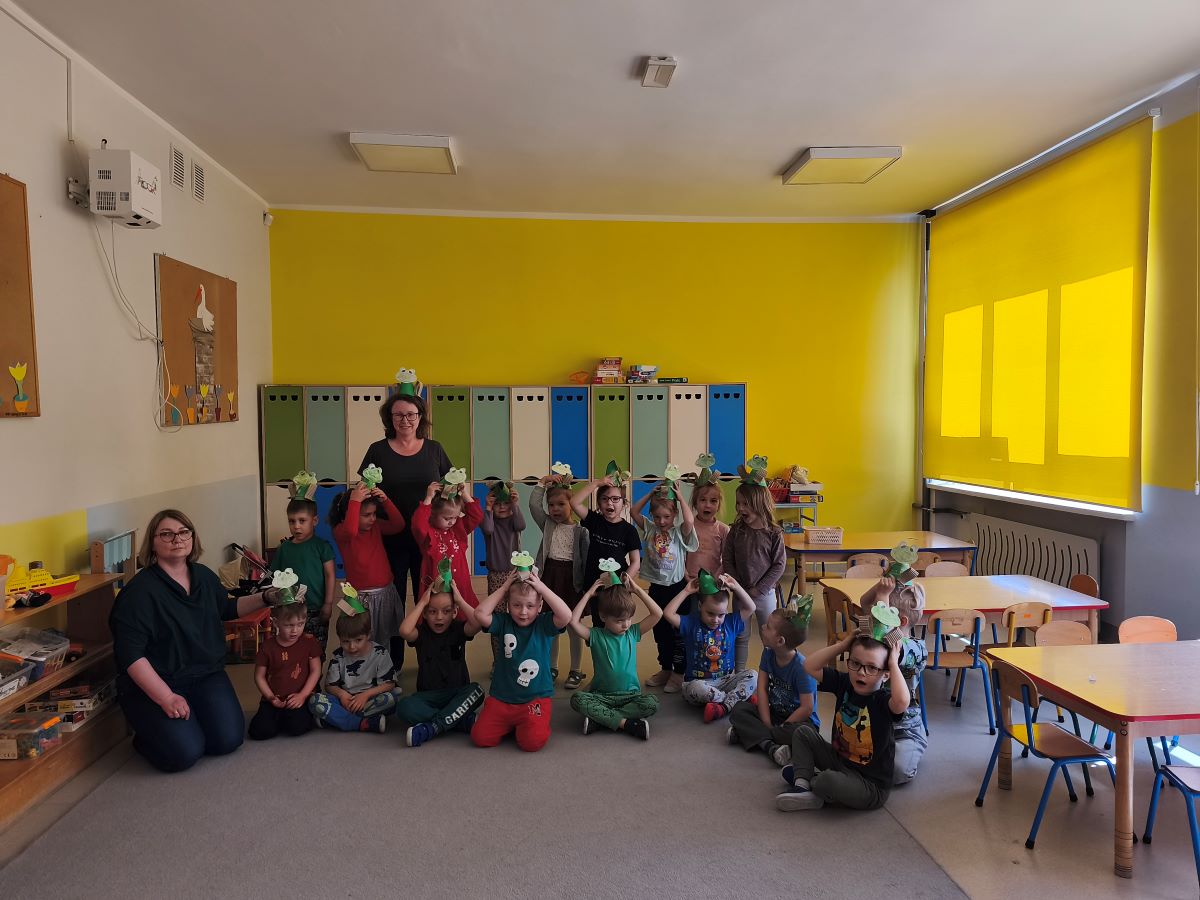 grupa dzieci przedszkolnych stoi razem w przedszkolnej Sali edukacyjnej i prezentuje wykonane przez siebie papierowe żabki. Dzieciom towarzyszy edukatorka ekologiczna i nauczycielka przedszkolna. Dzieci trzymają wykonane z papieru i tektury żabki na głowie.