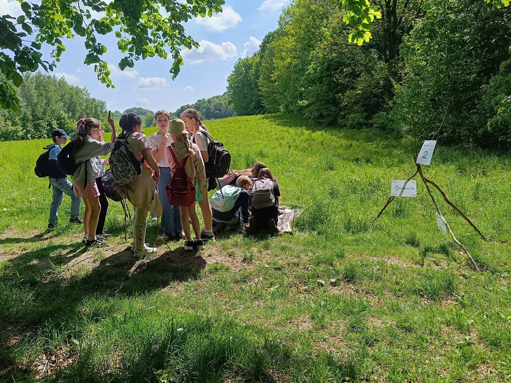 Zdjęcie przedstawia grupę dzieci z plecakami na zielonej polanie pod lasem w słonecznyc dzień. Część uczestników stoi w cieniu drzew, a pozostali siedzą na kocu i rozwiązują zadanie. Po prawej stronie widać konstrukcję z gałęzi z białą kartka i dużą czarną cyfrą 2 oraz plakat z nazwą Funduszowy Maj.
