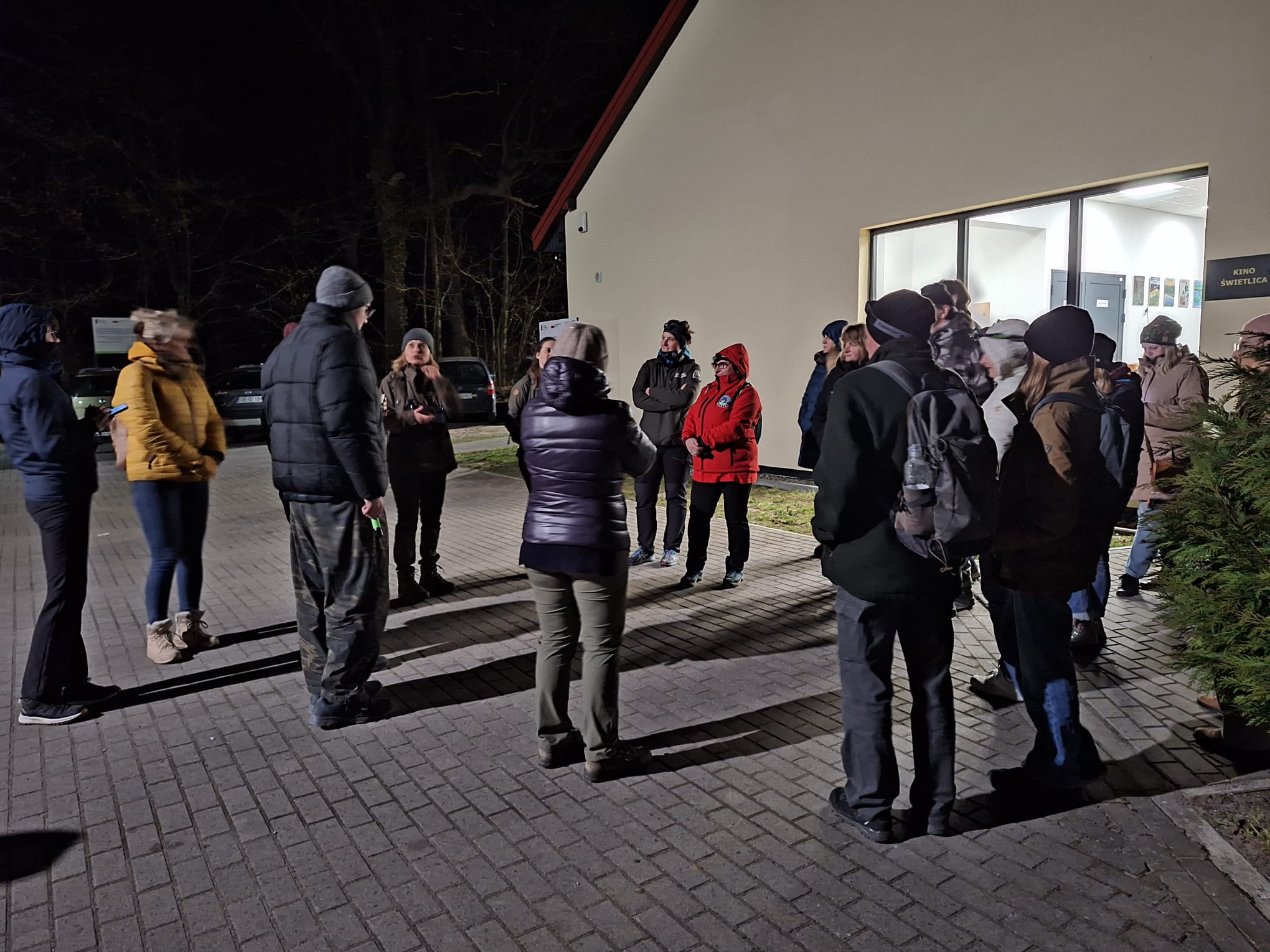 Grupa kilkunastu osób ubranych w zimowe kurtki, czapki i plecaki stoi wieczorem na brukowanym placu przed oświetlonym budynkiem. Uczestnicy ustawieni są w półokręgu i słuchają jednej osoby, która przemawia w centrum. W tle widać ciemny las oraz zaparkowane samochody, a światło z budynku.