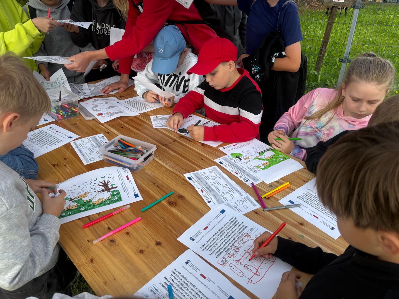 Dzieci siedzą przy drewnianym stole na świeżym powietrzu i rozwiązują zadania edukacyjne oraz kolorują obrazki z motywem owiec na polanie. Na stole leżą pojemniki z flamastrami i kredkami oraz arkusze papieru zawierające logotypy unijne i informacje o projekcie ochrony siedlisk w PK „Orlich Gniazd”.