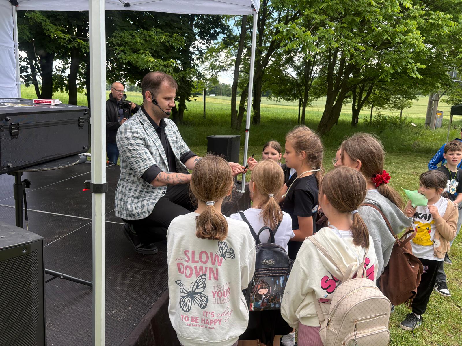 Pokaz iluzjonistyczny podczas plenerowego pikniku przy oddziale ZPKWŚ w Smoleniu. Mężczyzna w marynarce w kratę i z mikrofonem nagłownym kuca na krawędzi małej, czarnej sceny pod białym zadaszeniem, prezentując sztuczkę magiczną grupie zaciekawionych dzieci. Dzieci stoją tuż przed sceną na trawie i z bliska obserwują rekwizyt w dłoniach iluzjonisty. W tle, po lewej stronie, stoi mężczyzna z aparatem fotograficznym, a wokół rozciąga się zielony teren z gęstymi drzewami.