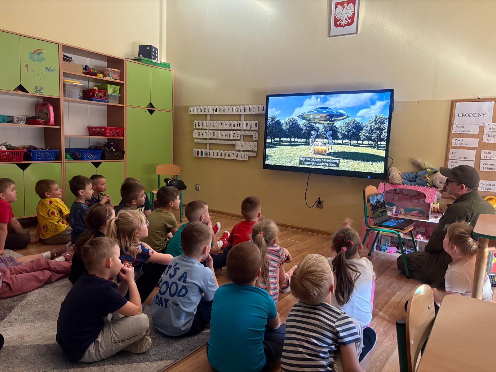 Dzieci siedzą na podłodze w sali przedszkolnej i oglądają na ekranie film edukacyjny. Zajęcia prowadzi mężczyzna siedzący obok z laptopem.